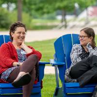 Two women sitting in blue lawn chairs outside talking with each other